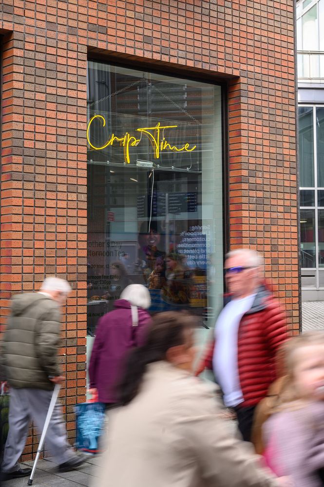 A group of people walk past a large glass window on a city street. The window belongs to a brick building and features a neon yellow sign reading "Crip Time." Reflections in the glass show more people and a display inside. The scene captures motion blur from the pedestrians, conveying a sense of bustling urban activity.