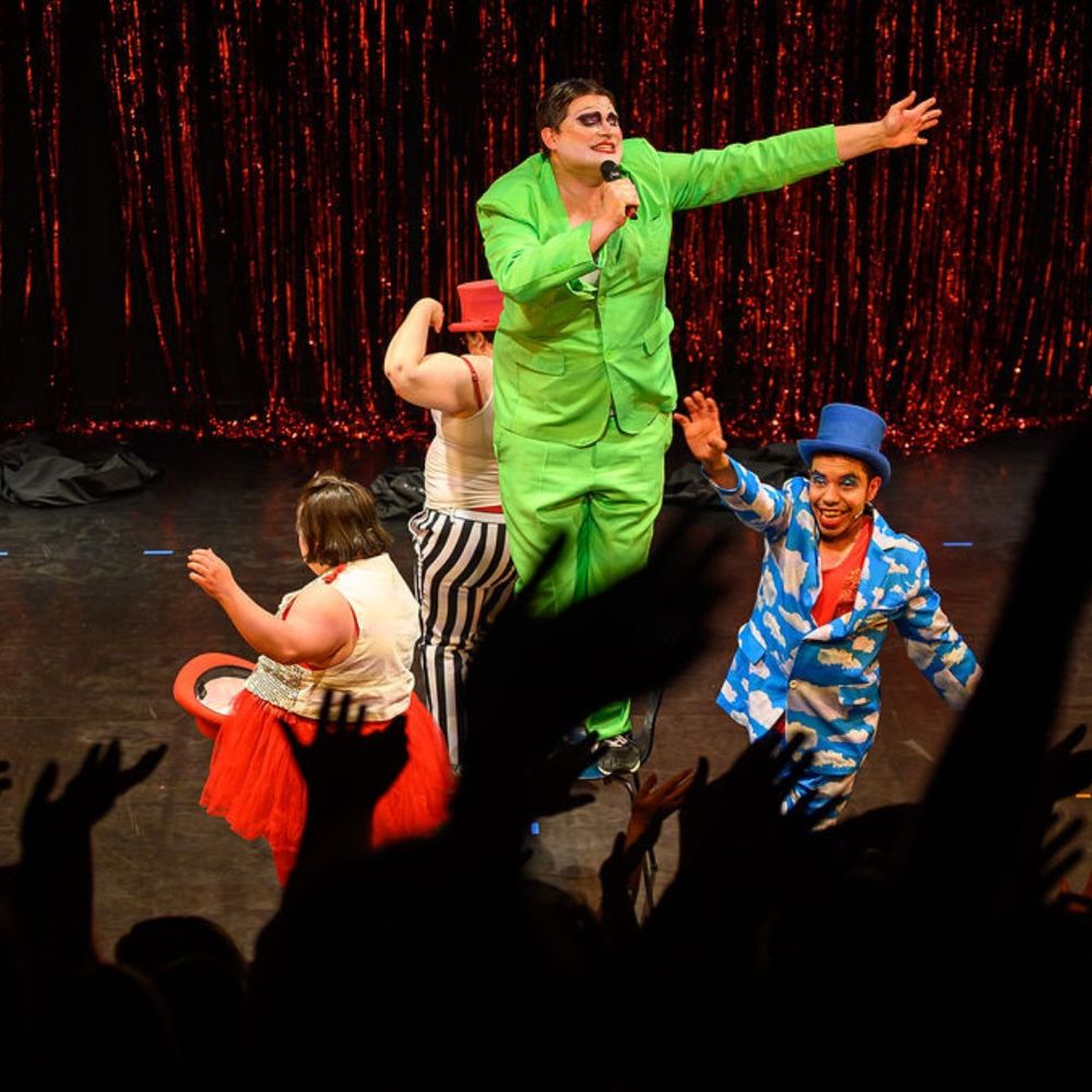 A lively stage performance featuring four performers in colorful, theatrical costumes. One performer in a bright green suit and dramatic face makeup sings into a microphone, reaching out energetically. Another performer in a blue suit with white cloud patterns and a blue top hat waves to the audience. Two more performers, one in a red tutu and another in black-and-white striped pants with a red top hat, also animate the scene. Hands of an excited audience are visible in the foreground.