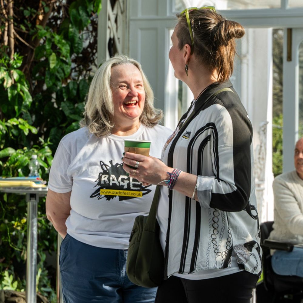 Two women stand smiling and chatting in a bright indoor space with greenery. The woman on the left, with gray hair, wears a white t-shirt with the word “RAGE” and the DaDaFest website printed on it. The woman on the right, with short brown hair and green sunglasses on her head, wears a black and white striped jacket and holds a green paper cup. They appear to be enjoying each other's company.