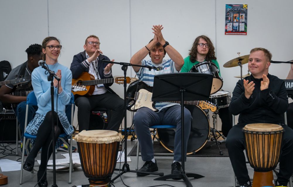 A group of young disabled musicians are seated behind music stands and microphones. some of them have drums next to them. One sits in front of a full drum kit and a few have electric or acoustic guitars. They are all clapping and smiling.
