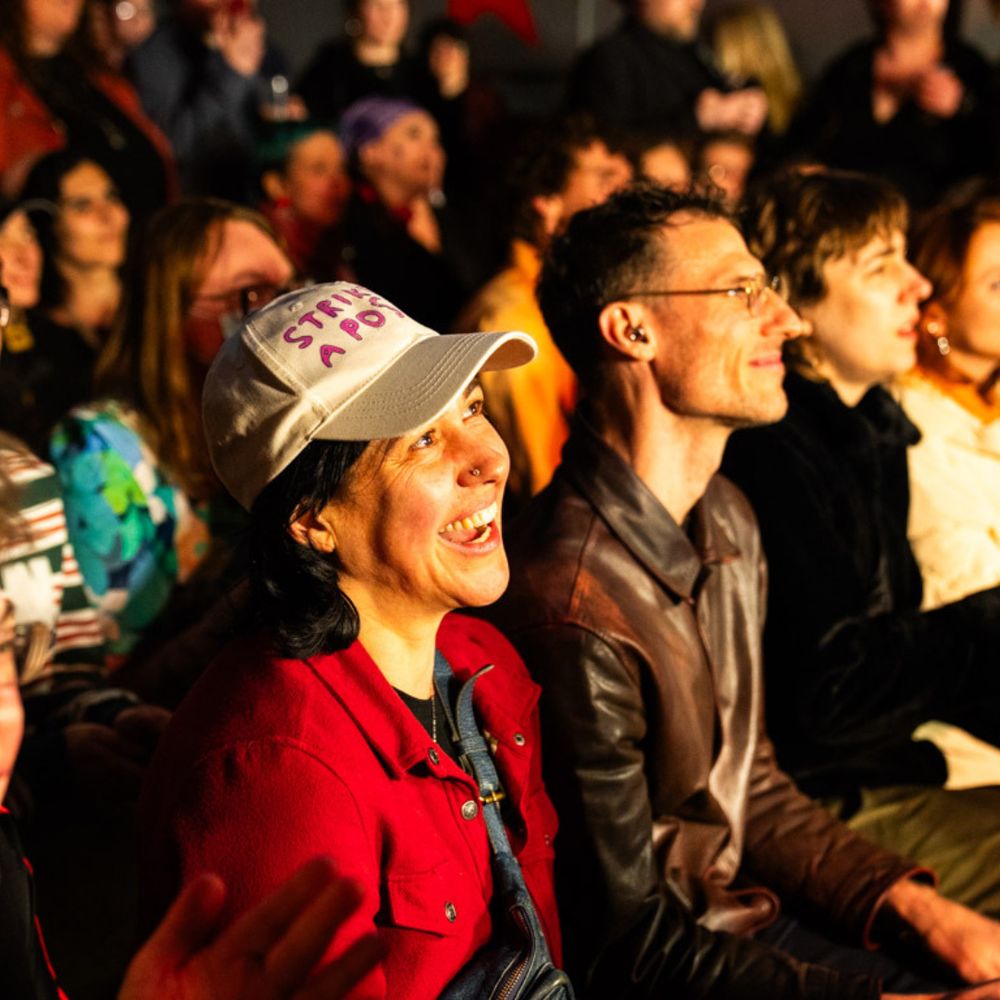 A lively and diverse audience watches an event with visible joy and engagement. In the foreground, a person wearing a white cap with handwritten text “STRIKE A PO…” (partially obscured) and a red jacket is smiling brightly. Next to them, another person in a brown leather jacket also appears to be enjoying the performance. The background is filled with people, some clapping, all focused on the stage out of view.
