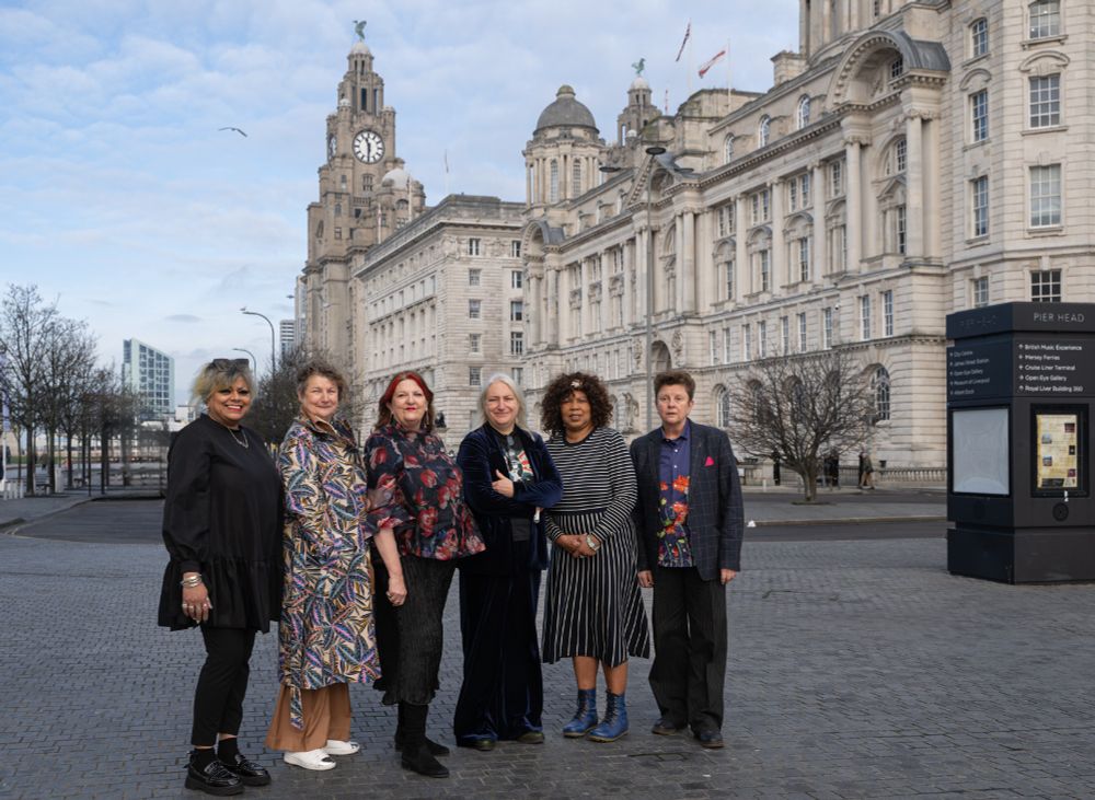A group of people stand side by side, smiling into the camera. They are about to speak at the Rushton Lecture, for DaDaFest. The panellists are: Kaite O'Reily, Julie McNamara, Cheryl Martin and Ruth Fabby. They are dressed formally, they are stood outside in front of the iconic Liver Building, seen in the distance. Blue skies feature.