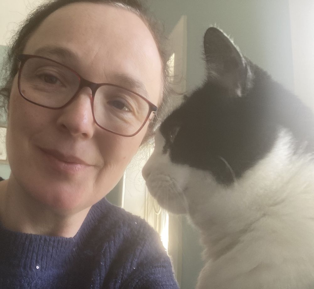 Professor Jo Brück, a dark haired woman with glasses and a blue sweater & her cat Willow, a black & white cat, looking at her 