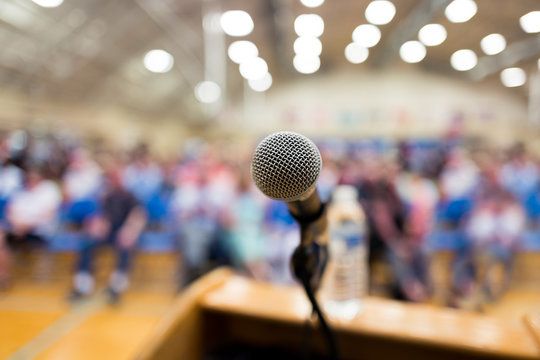 Picture of a microphone at a podium with a blurred audience in the background.