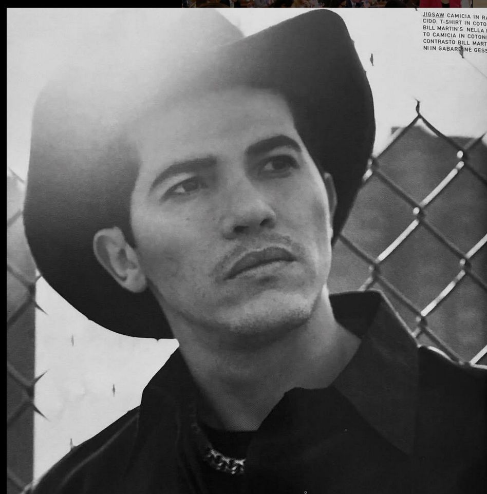 black-and-white shot of John Leguizamo looking unbearably hot, statuesque-but-soulful, wearing a cowboy hat and a thoughtful expression