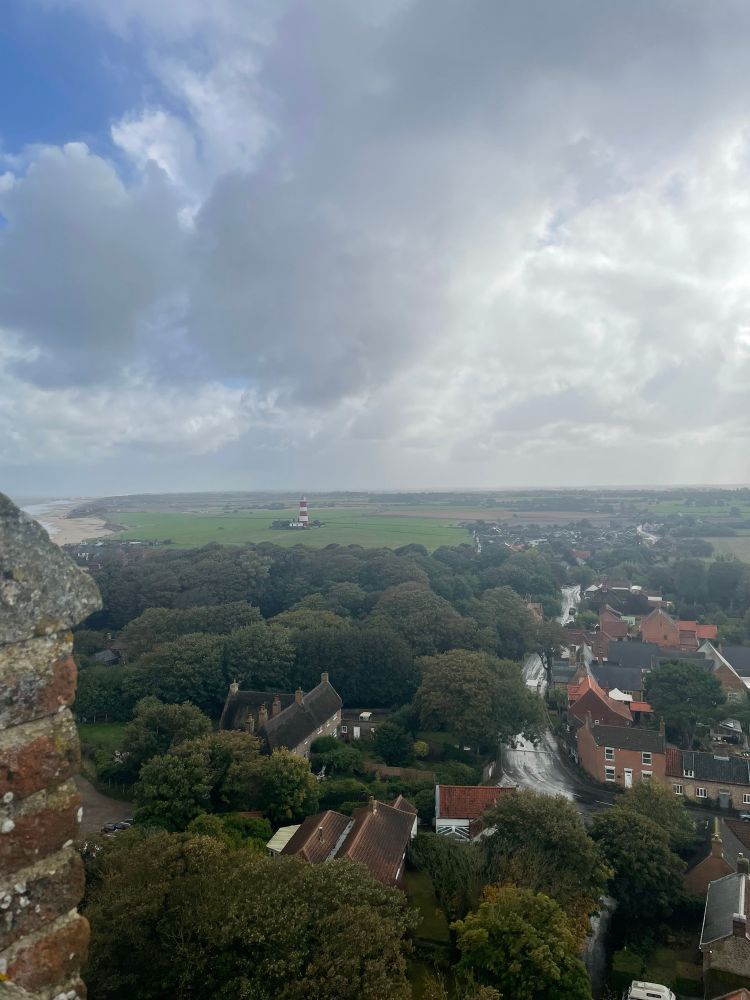 Looking south. View of the main street in Happisburgh from the church tower. Mostly trees, a few houses, a bit of beach and sea and the red and white striped lighthouse.