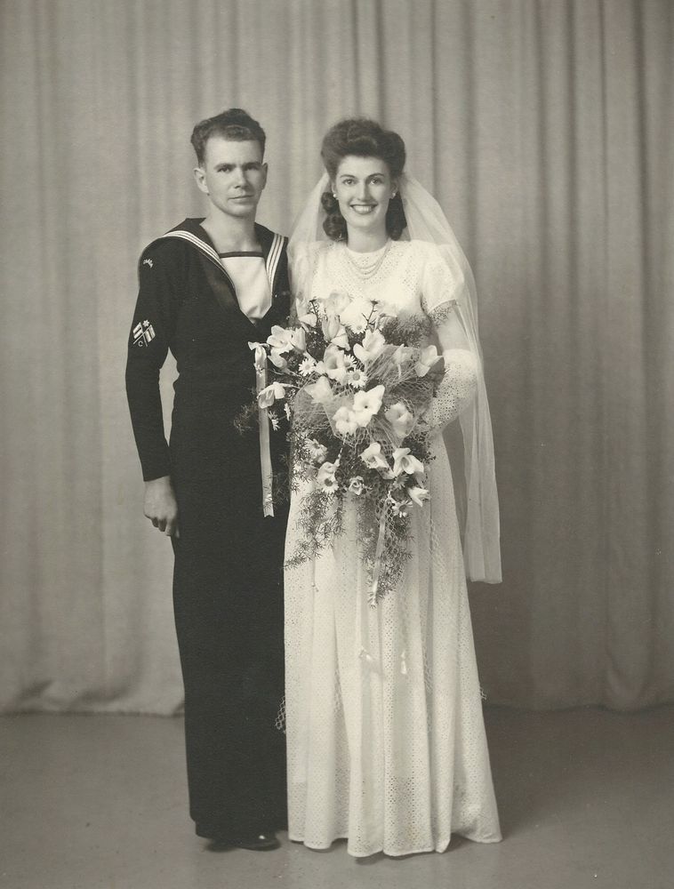 A black and white photo of a bride and groom from 1945. The groom is in his dress blues naval uniform and the bridge is wearing a floor length white gown, mid-length veil and carrying a large bouquet. Her hair is style beautifully and she is wearing pearl earrings and a pearl necklace, with a big smile on her face. 