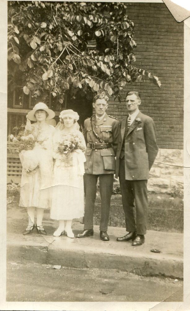 A grainy black and white photo of a wedding couple in 1925 and their best man and maid of honour. The groom is dressed in his military uniform and the bride is wearing a white, ankle-length dress and a hat with a lace veil. She and her maid of honour, also dressed in white, are both holding large bouquets. The best man is wearing a suit with a striped tie. Both men are standing with their hands clasped behind their backs, straight posture. 