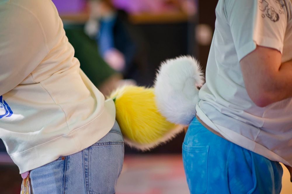 2 people facing butt to butt wearing short nub tails. The person on the left is wearing a yellow tail and the person on the right is wearing a white bunny tail. They are touching.