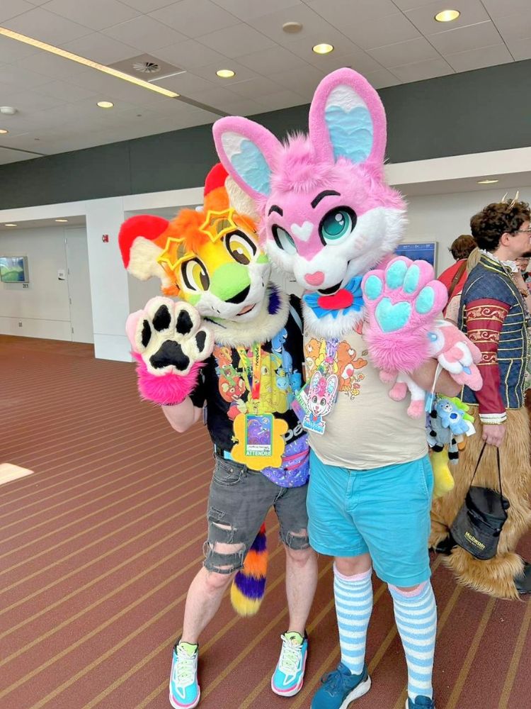 Bryte's rainbow plushie kougra neopet partial fursuit and me in my pink bunny Sugarbun Fursuit both waving to the camera. Though I am awkwardly looking slightly away from the camera cause I instinctively turned my face to camera rather than my actual fursuit head lol.