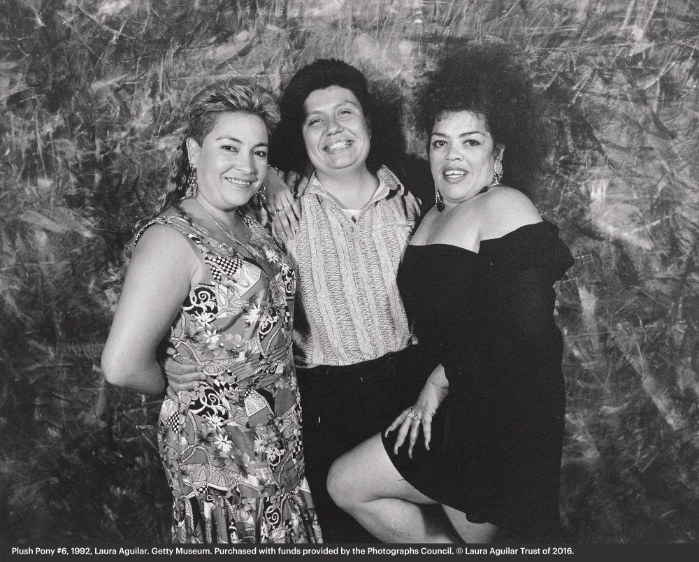 Plush Pony #6. Portrait of three woman dressed in club outfits who are implied to be lesbians. The first and third woman are wearing dresses, but the second woman in the middle is wearing a polo and slacks. They are posed closely together and smiling indicating they have a friendly bond between them. They are in front of a typical photo studio backdrop, but are inside of the Plush Pony lesbian bar.