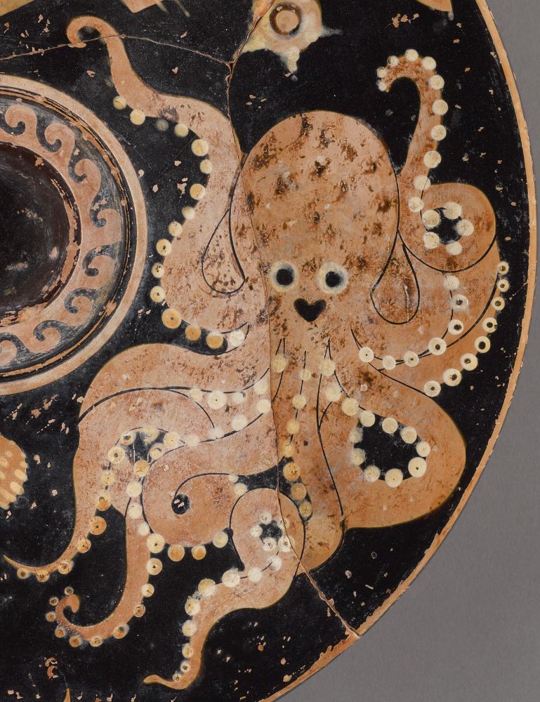 Close up on an ancient plate featuring a light pink octopus with bright eyes and a smile. The octopus has two arms waving above its head in a friendly manner. 