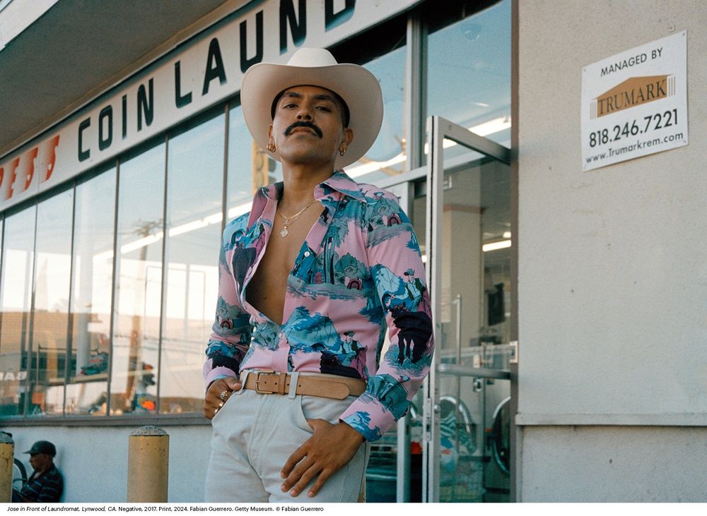 A model, José, poses in front of a laundromat in Lynwood, California. A Mexican-American man, he is dressed in the style of a modern cowboy. He wears a white cowboy hat, a leather belt, and a button-up shirt with a print that features a cowboy lassoing a cow. However, there are elements of the outfit that challenge queer assumptions of how a cowboy, or ranchero, dresses. Namely, the shirt is mostly pink and teal- a bright colorway that isn't typical of most cowboy getups. Secondly, José wears several pieces of jewelry: multiple hoop piercings in his ears, two or more medallion and chain necklaces, and three rings on his right hand. He leans confidently against a concrete post in front of the entrance to the laundromat, gazing down at the viewer in confident defiance. 