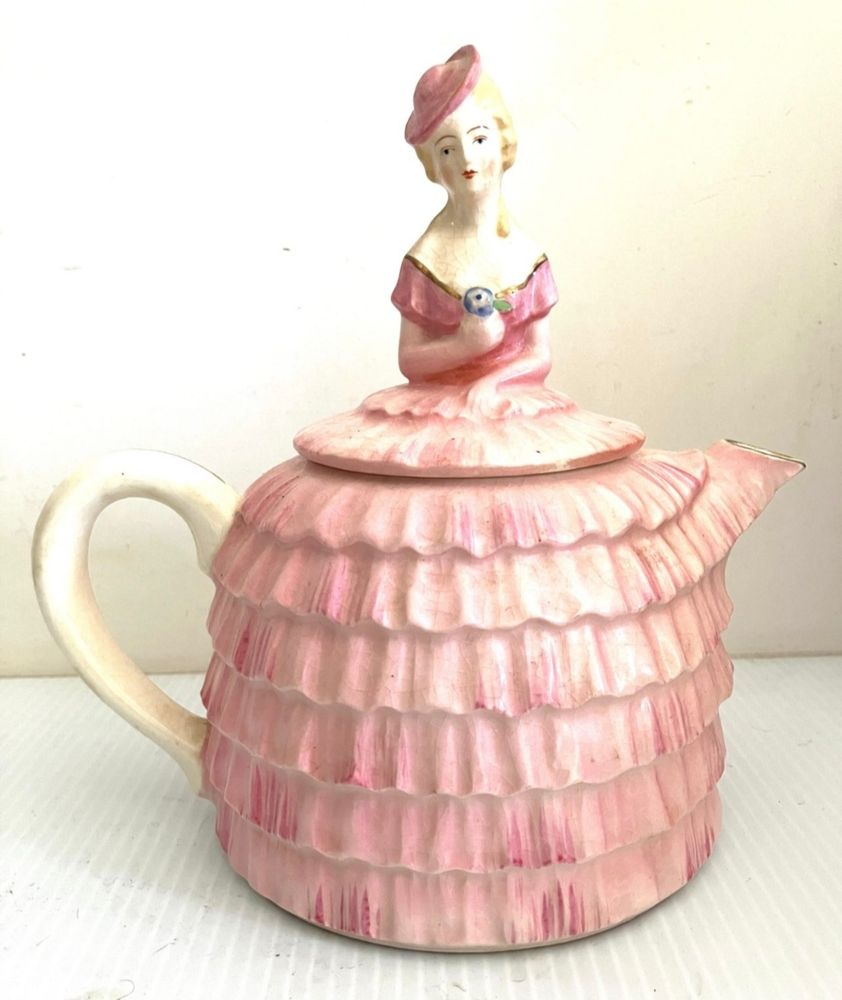 A beautiful teapot. The pot is a pale pink, 6-tiered skirt. The top is a woman's torso in the top of the pink dress. It's off-the-shoulder. She had blond hair and a matching pink hat on a jaunty angle