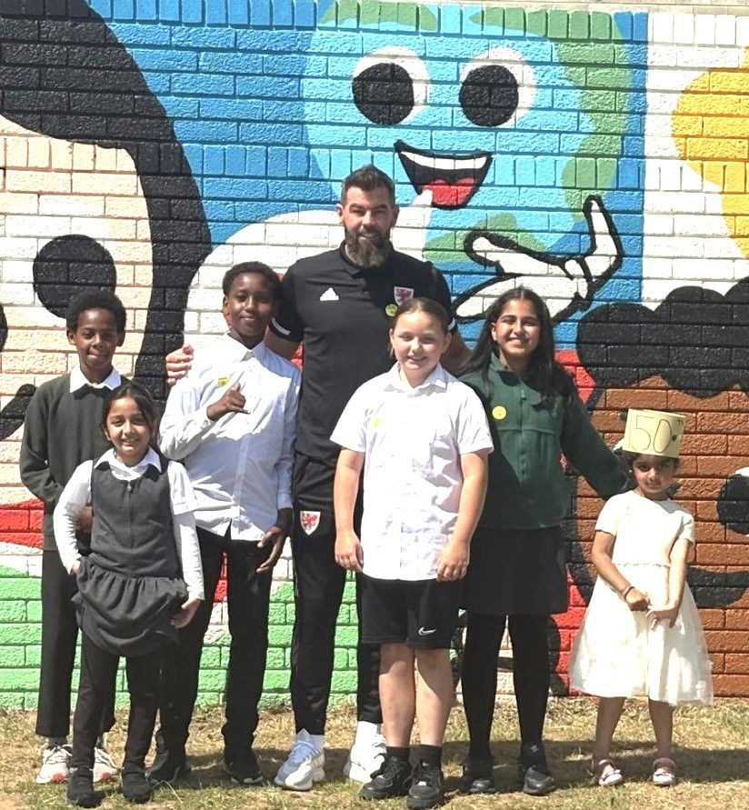 Mae grŵp o ddisgyblion ysgol yn sefyll, gydag oedolyn o flaen murlun, ar wal.