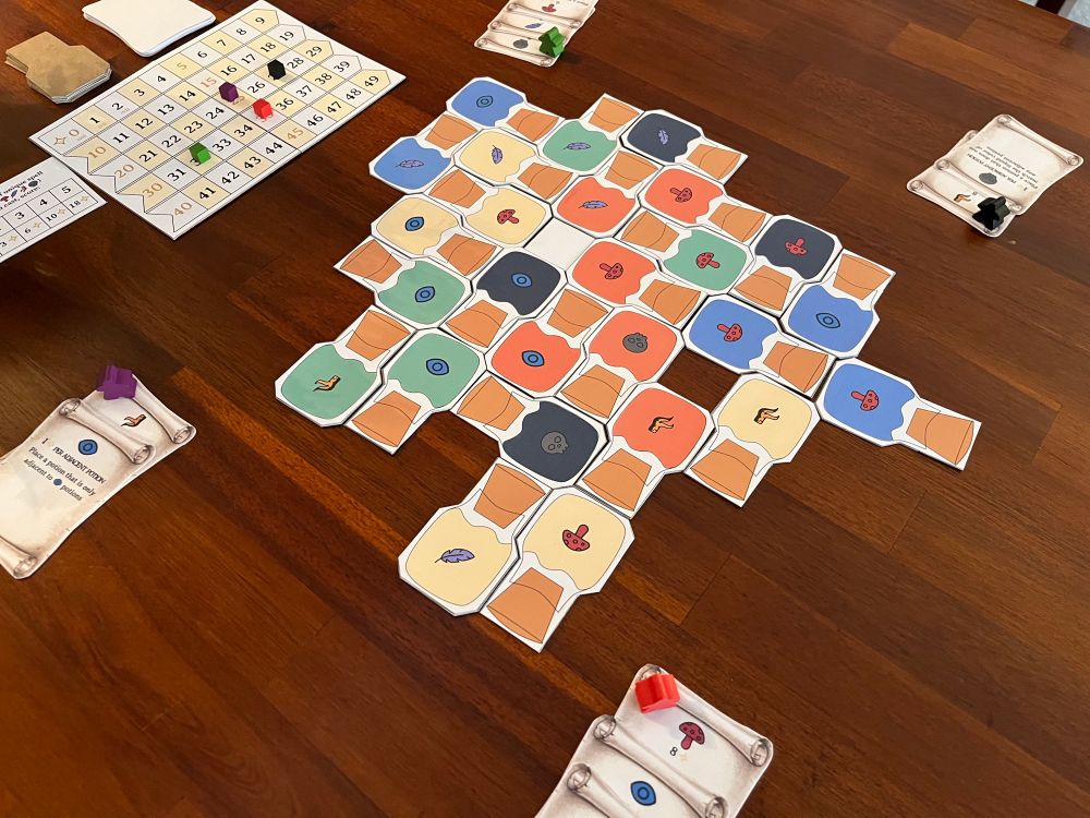 Potion shaped tiles aligned together with different colors and symbols on them. Nearby is a scoreboard with cubes on top and some stacks of spell cards.