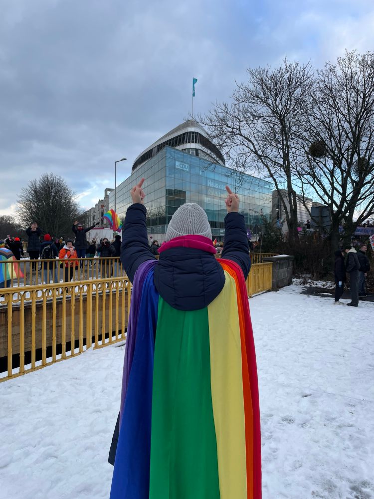 Person mit Regenbogen Umhang steht vor der cdu zentrale mit dem Rücken zum Foto und hält beide Mittelfinger hoch