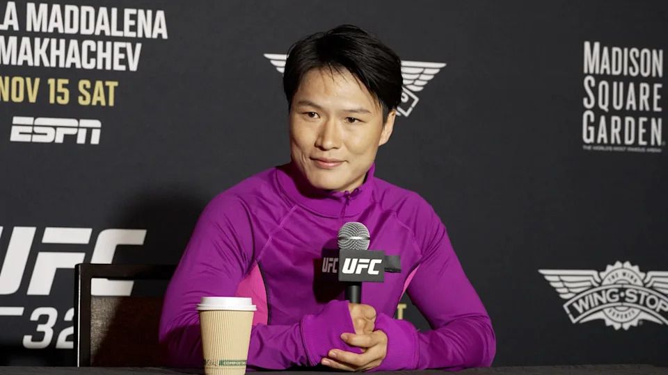 zhang weili wearing a purple top sitting in front of UFC backboard smiling confidently 