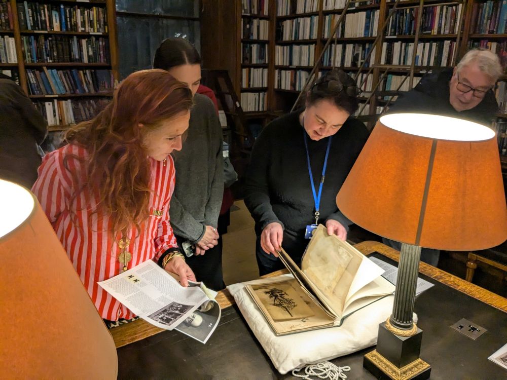 An archivist is turning the pages of a scrapbook/photograph album which supported on a book rest on a table in a library. She is surrounded by visitors looking at the album. As always, the overly large shades of the table lamps of the RAS library are in the foreground. 