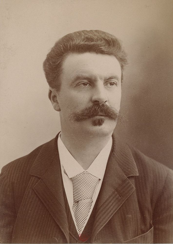 A sepia-toned photo portrait of Guy de Maupassant. His dark hair is brushed back, his mustache is fuller and more unruly than a pillow case stuffed with cats and he has a soul patch. He's dressed formally: white collared shirt, neck tie, suit coat and vest.
Image source: Gallica, the Digital Library of France
