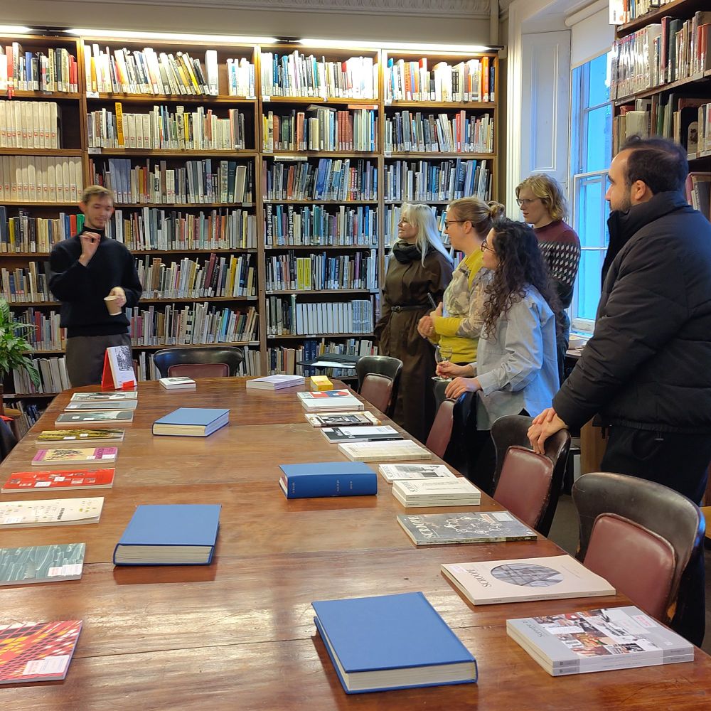 A colour photo of Nicholas Frayne, one of the Scroope journal editors, talking to the audience that gathered around the table. On the foreground the past issues of Scroope are visible, lying on the long table.