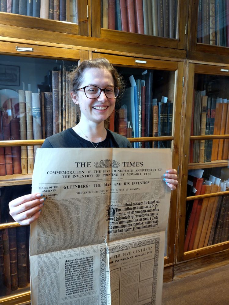 A photo of Victoria standing in the AHA Library Rare Books room, holding The Times newspaper from 1940 in her hands and smiling.