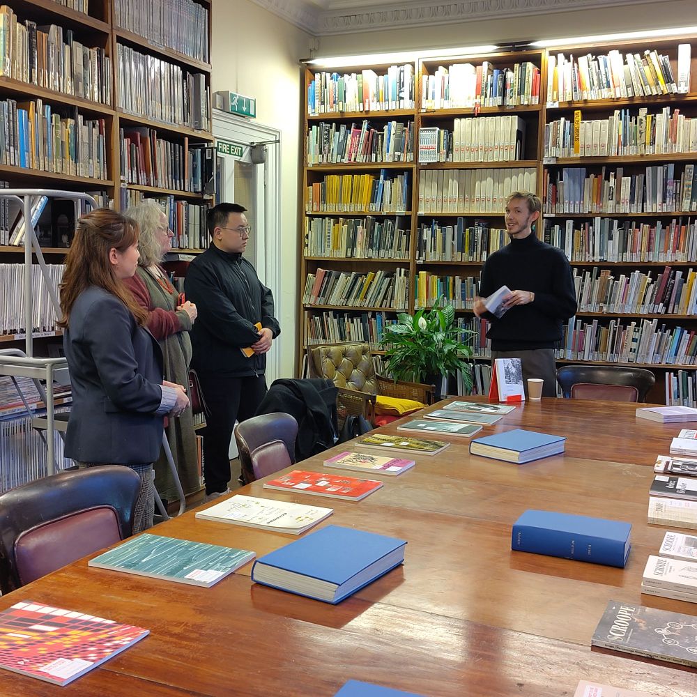 A colour photo of Nicholas Frayne, one of the Scroope journal editors, talking to the audience that gathered around the table. On the foreground the past issues of Scroope are visible, lying on the long table.