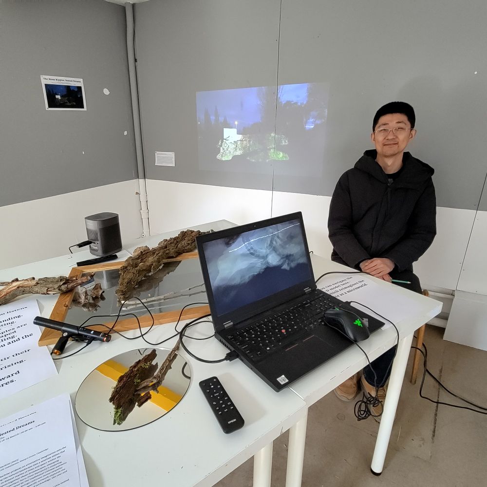 A colour photo of Lingzheng Zhu's installation at the AHA Faculty gallery. A laptop with software that changes voice tones into visual patterns and words and displaying them is in the foreground, on the table. Next to the laptop, there are mirrors and wood bark taken from he Sheep's Green common grounds. In the background, a film made by Zhu at the Sheep's Green common grounds is projected on the wall. The installation author is smiling and sitting on the chair next to the exhibition.
