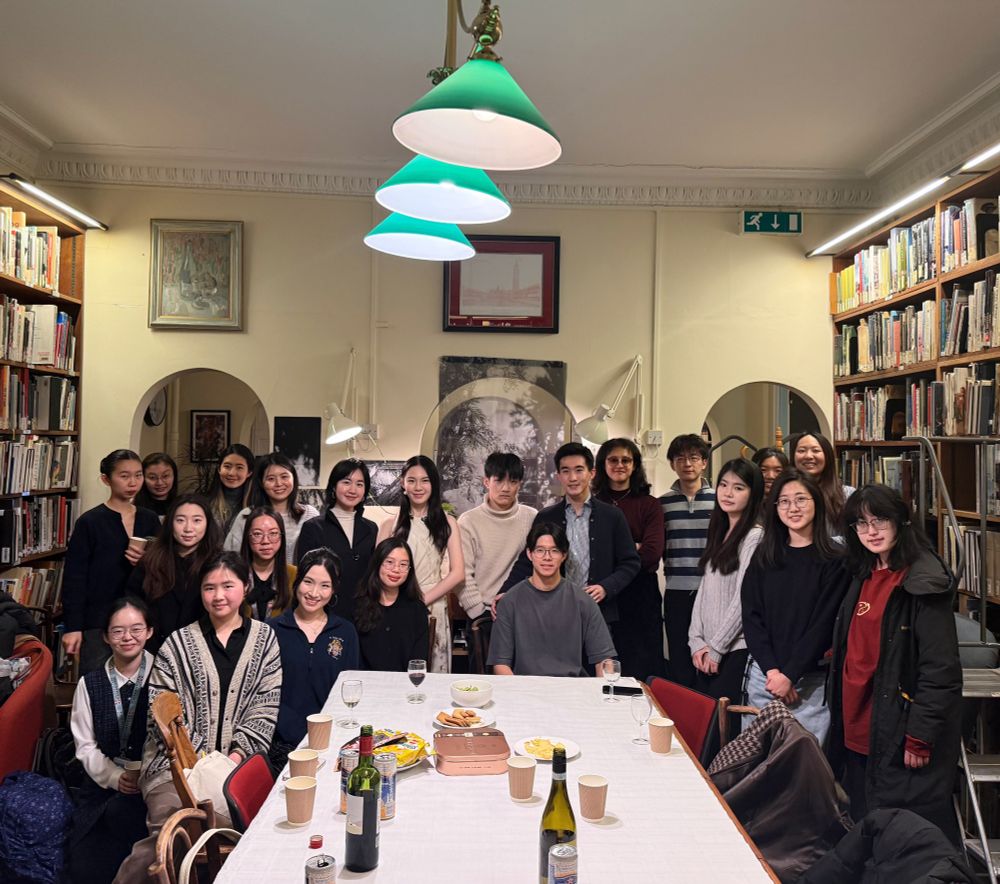 A group photo of the visitors of the exhibition in the library History of Art Room, with the artist in the centre and 20 people around, sitting and standing. On the foreground there is a long table with drinks and snacks, and the exhibition is behind the group.