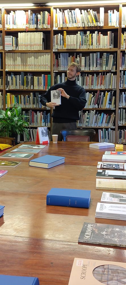 A colour photo of Nicholas Frayne, one of the Scroope journal editors, standing with the new issue of Scroope in his hands. On the foreground the past issues of Scroope are visible, lying on the long table.