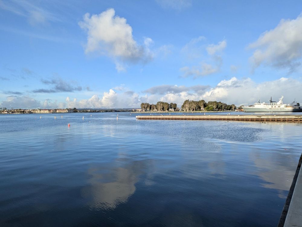 water view with clouds and a boat, dock, and trees in the distance.