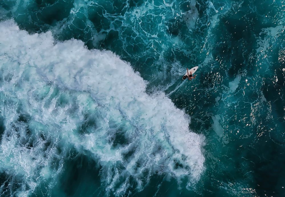 Aerial view of a lone surfer on a white surfboard, riding a powerful wave in El Tunco, El Salvador's turquoise waters. White foamy crest contrasts with the deep blues of the Pacific Ocean. 
