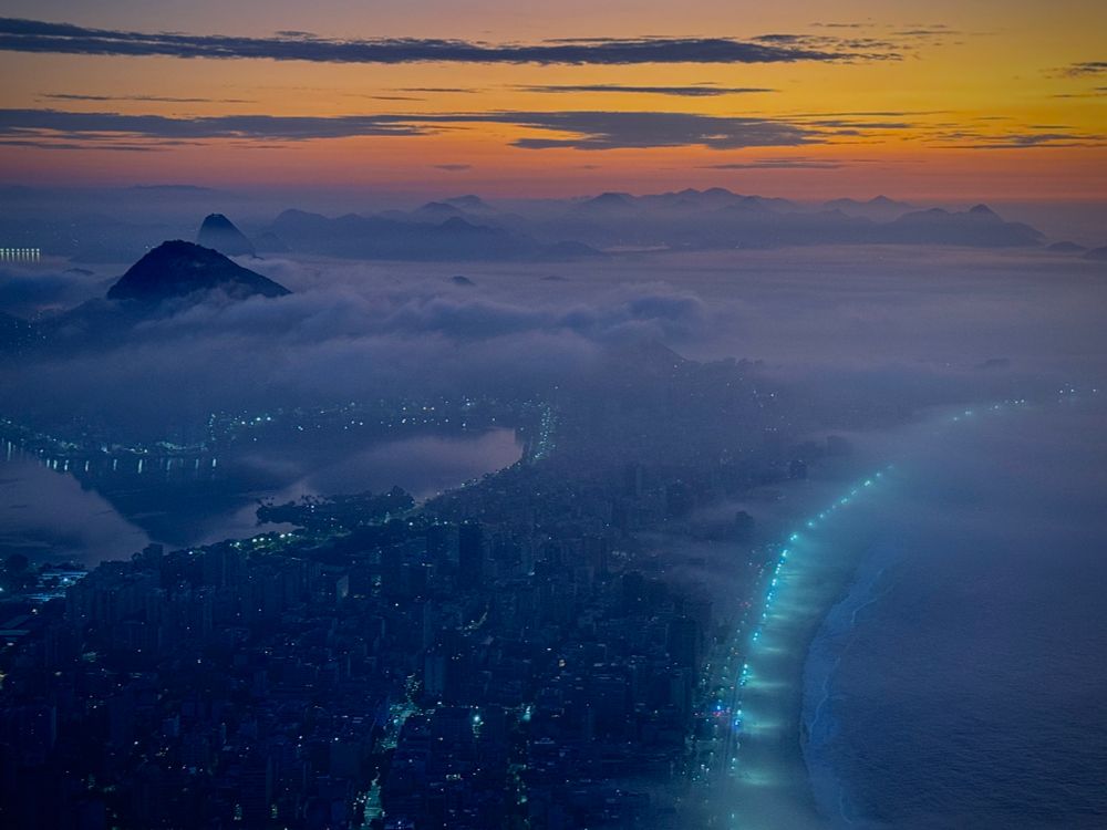 First light embraces Rio de Janeiro, a city cloaked in the gentle morning mist. City lights twinkle along the gentle arc of Ipanema beach. The sky hints at sunrise colours as iconic mountains peek through the ethereal fog. 