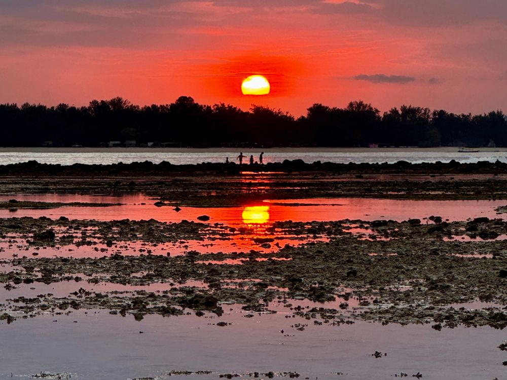 From a beach on Gili Air, the glowing red sun sinks into the horizon, its reflection shimmering in tide pools as silhouetted children wander through the rocky shallows.