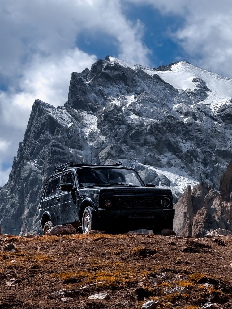 A lone black Lada Niva sits at the edge of a rocky slope in the Tien Shan mountain range, dwarfed by the immense, snow-draped peaks rising sharply under a shifting sky.