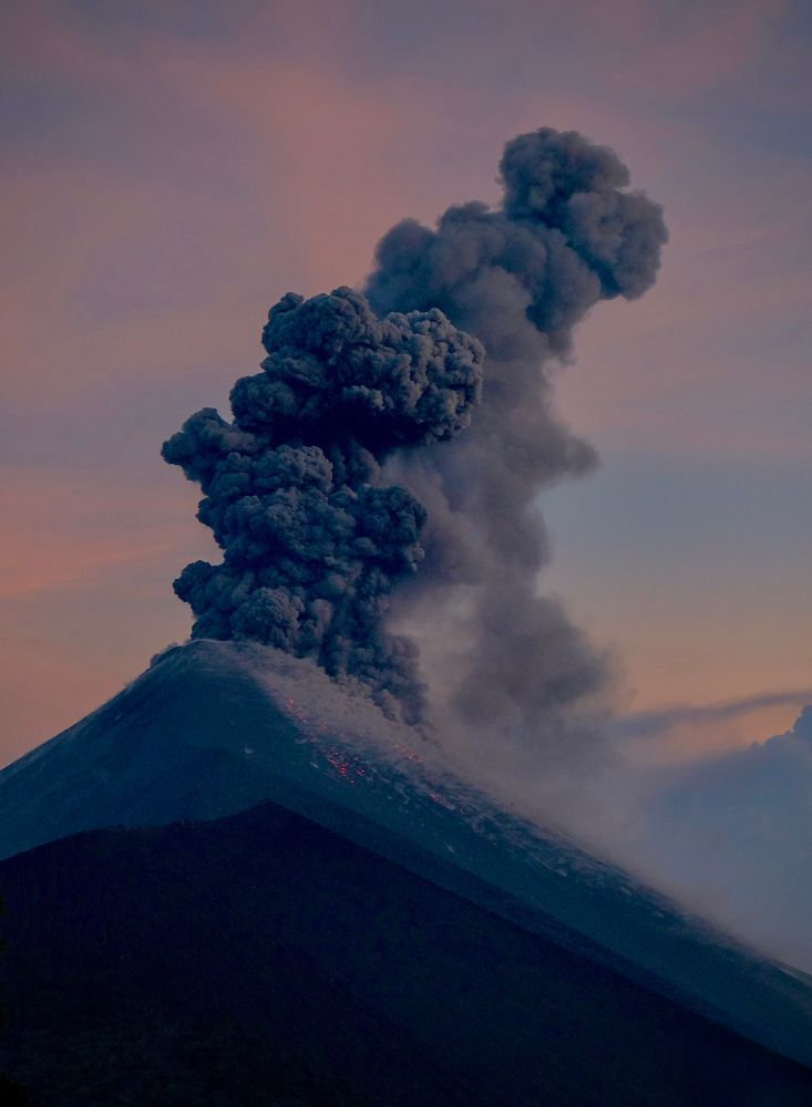 Just outside Antigua, Guatemala, Volcán de Fuego erupts, spewing a thick plume of dark ash into the soft, orange and blue of the evening sky. Glowing lava cascades down its steep slopes in a dramatic volcanic display.