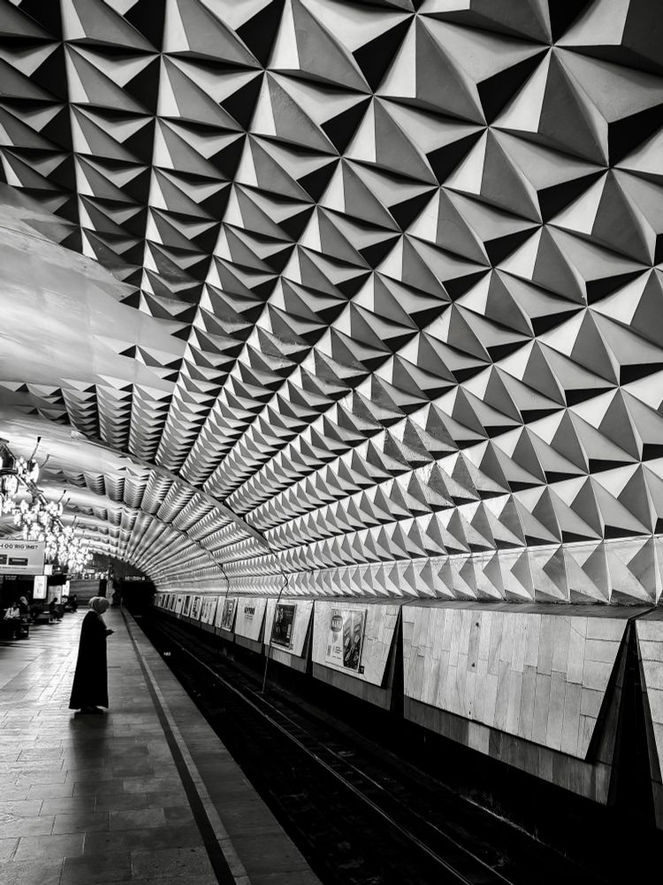 A black and white photo of the geometric, faceted ceiling of Beruniy Metro Station in Tashkent, stretching above a quiet platform where a lone person stands near the tracks under dramatic, repeating pyramidal ceiling panels.
