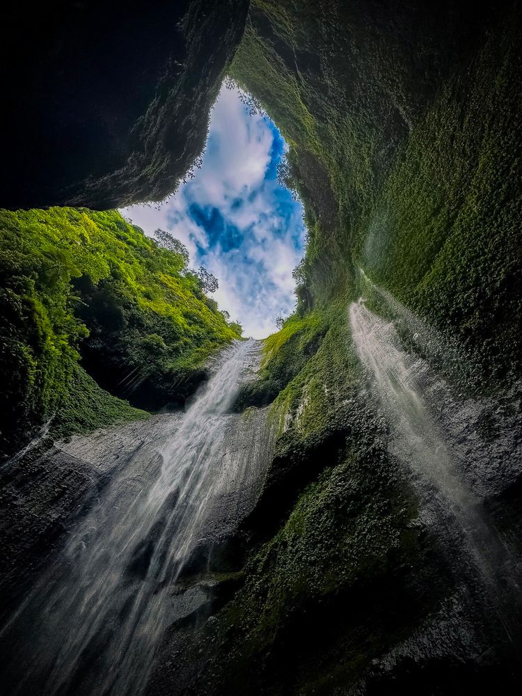 Seen from the base, a towering waterfall plunges 200 meters down a canyon wall, vanishing into soft mist at the foot of a cathedral of stone and light. Morning sunlight grazes the cliff walls, draped in moss and lush vegetation.