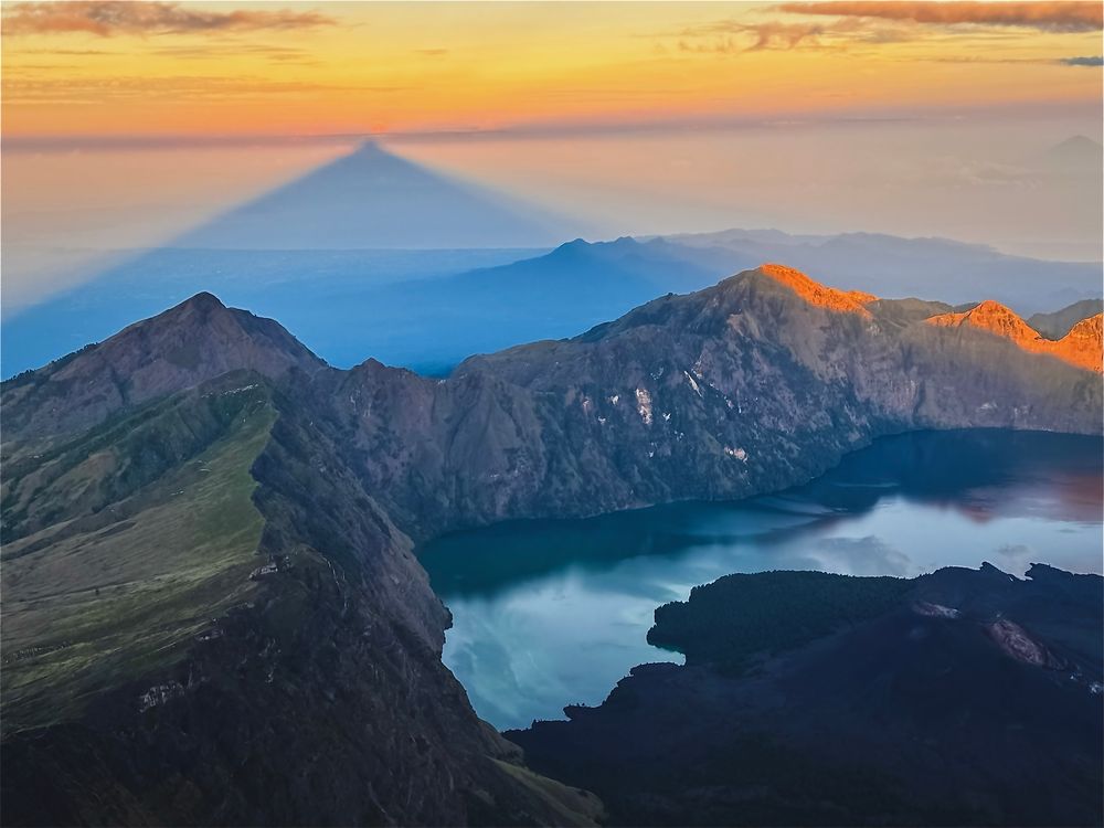 From the summit of Mt. Rinjani on the Indonesian island of Lombok, the rising sun paints the ridges gold while the mountain’s shadow pierces the horizon, forming a fleeting pyramid in the sky. A still, blue crater lake and a smaller volcano can be seen below.
