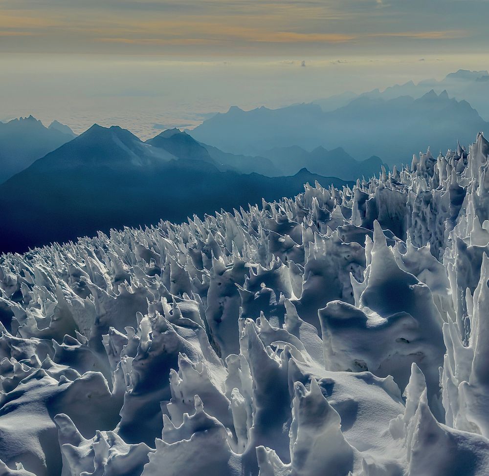 Surreal landscape of spiky white ice formations cover the slopes of Huayna Potosí mountain in the foreground, looking like frozen waves or peaks of meringue. Misty blue mountains can be seen in the background.