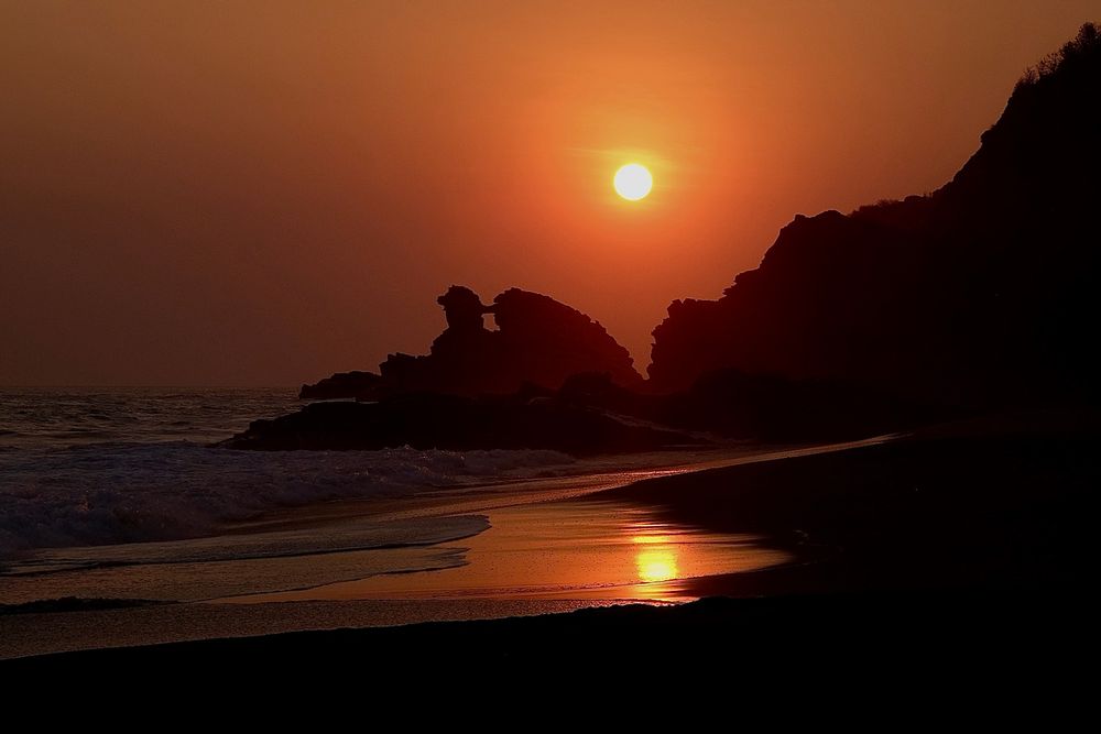 A vivid orange sunset over Playa Mermejita in Mazunte. Silhouetted rock formations along the coast resemble a human profile gazing out to the ocean. Gentle waves lap the shore, reflecting warm light across the wet sand.
