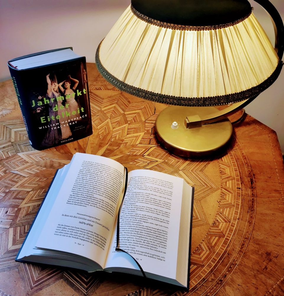 Auf einem gemusterten Holztisch liegt unter einer Lampe das aufgeschlagene Buch "Jahrmarkt der Eitelkeit", dahinter steht ein weiteres Exemplar des Buchs.