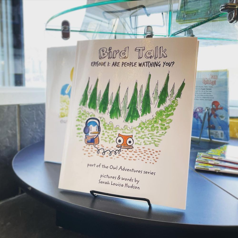 A photo of the picture book "Bird Talk" on a stand in a bookstore. The book's cover shows Owl and Penguin in the woods wearing sweaters and recording their podcast.