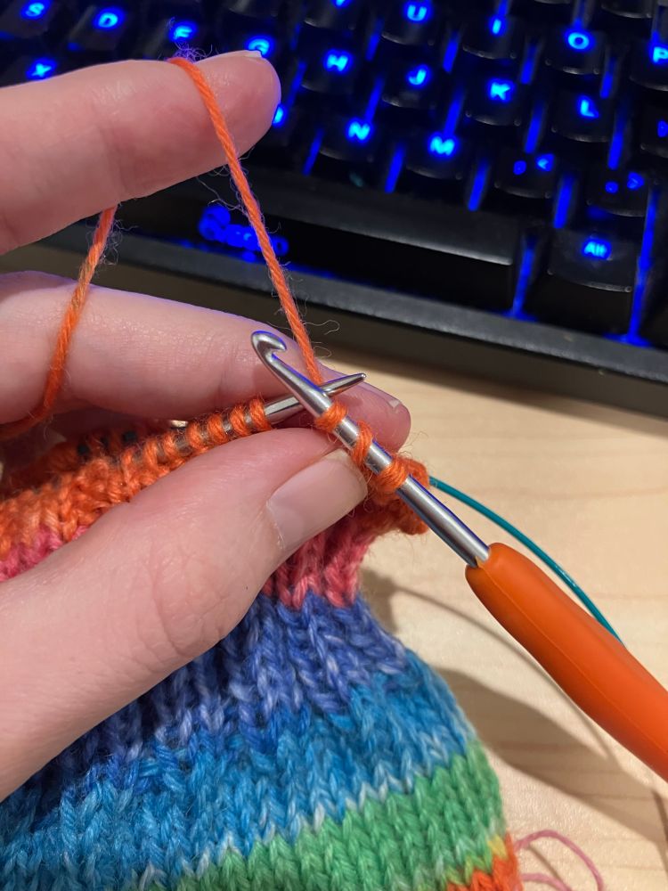 Some knitting is visible. Most of the knitting is one a knitting needle but instead of a second needle there is a crochet hook on the right. Three stitches (actually it is one stitch, a yarn over, and another stitch but you can’t tell that from the photo) are sitting on a crochet hook.