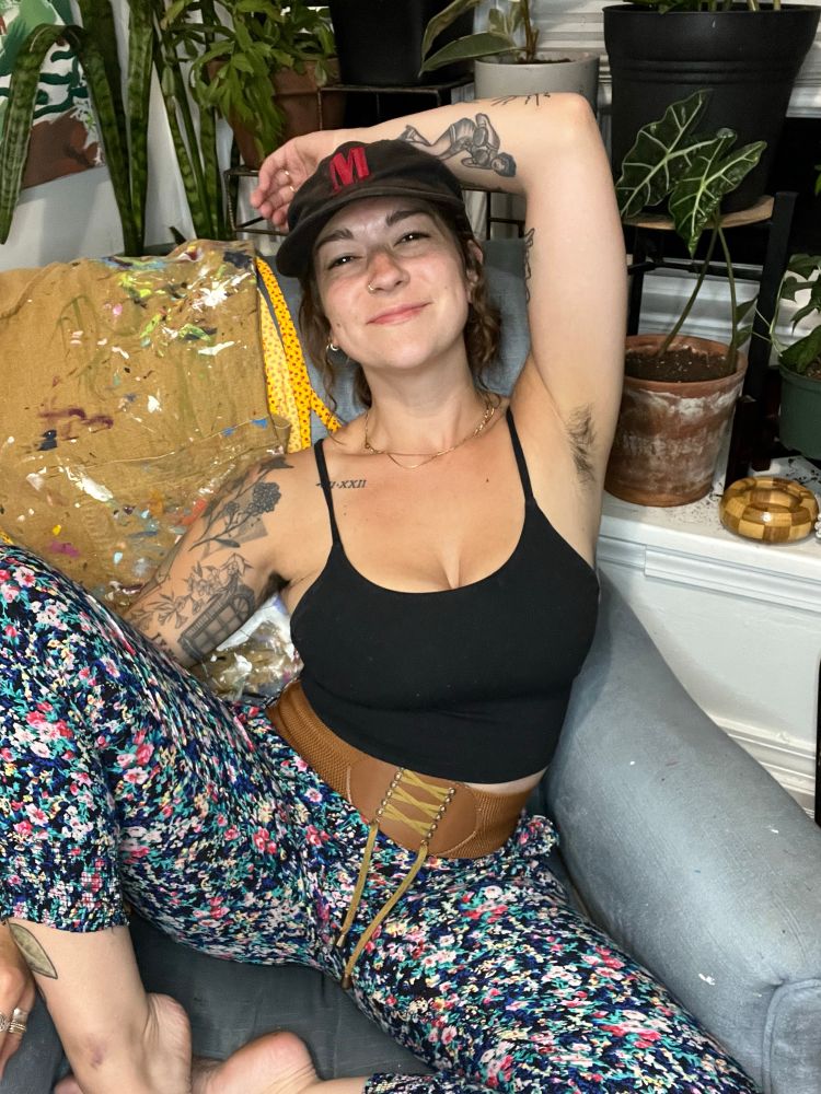 Trouble in her element: luring you in with her pretty, dark, captivating eyes. She’s in her painting chair, on the studio, surrounded by plants. She’s sitting with her legs wide open, thin floral pants on the bottom, thin strapped black cami on top- covering her perfect, natural tits. She’s tattooed, hairy, and confident. Arm above her cute, Marlboro-hat adorned head, she’s smirking at you, knowing that you’ll stop for a second to look. That’s all she needs