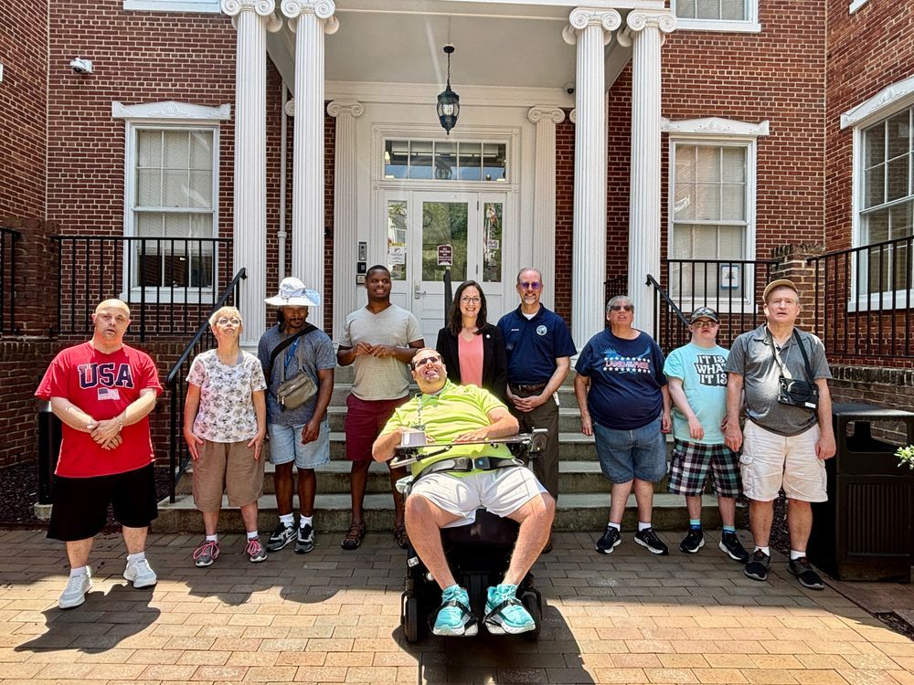 Jessica Fitzwater [standing in a neon pink blouse and black blazer] and Mayor O'Connor [left, wearing a navy polo] standing in front of Winchester Hall with individuals with disabilities.