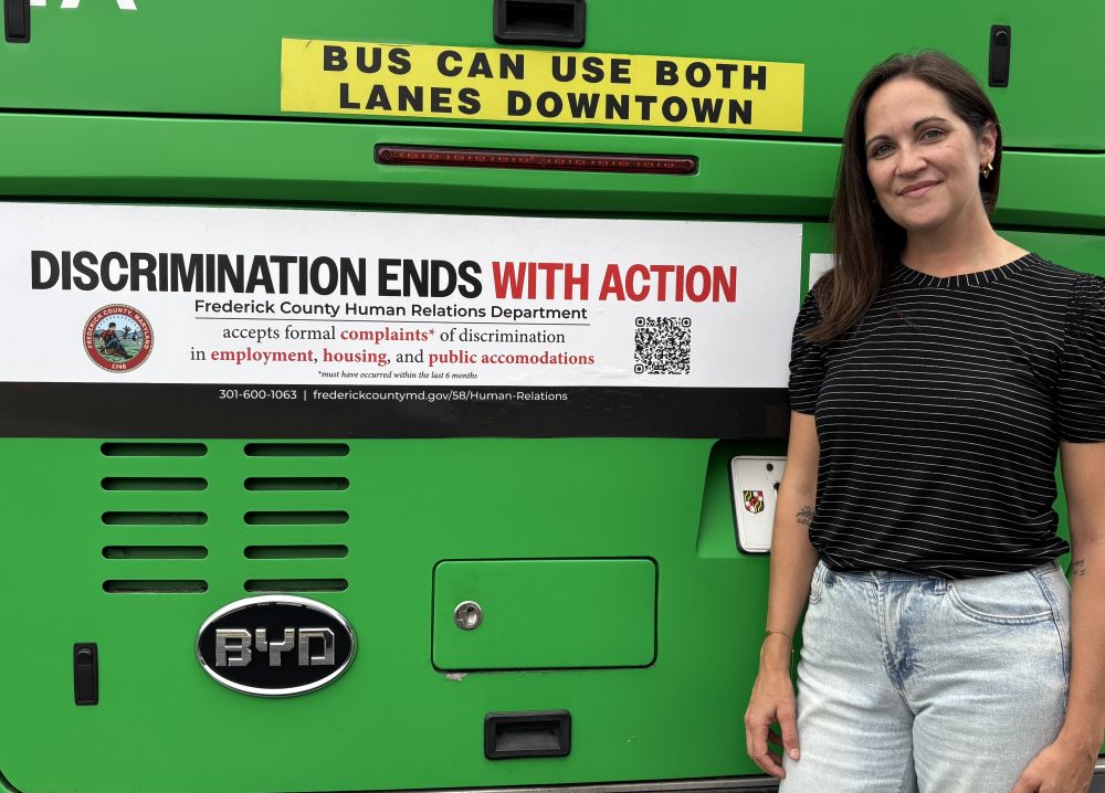 Jessica Fitzwater in a black and white striped shirt with jeans [right] standing next to a Frederick County green transit bus with a sign that says "Discrimination ends with action" by the Frederick County Human Relations Department. It accepts formal complaints [made within the past 6 months] of discrimination in employment, housing, and public accommodations. Website: frederickcountymd.gov/58/Human-Relations