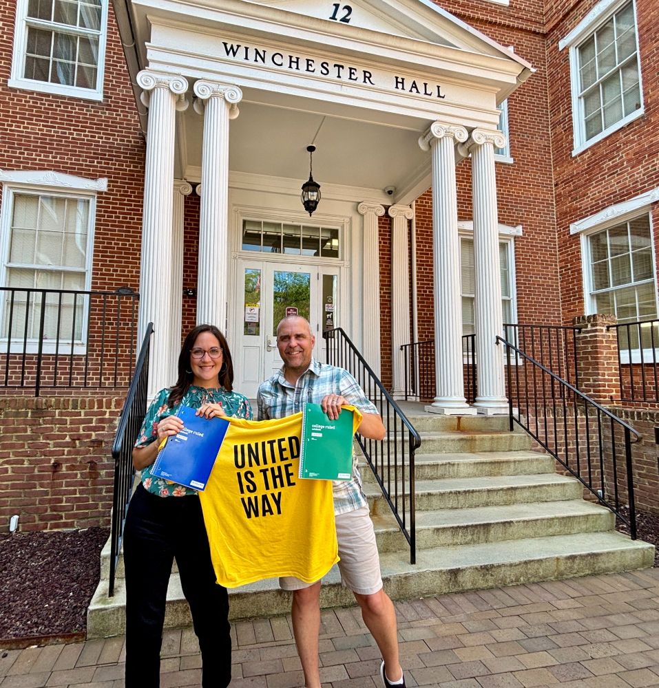 Standing in front of a brick Winchester Hall, Jessica Fitzwater [left] is holding a blue notebook and standing next to Ken Oldham [right], President & CEO of United Way Frederick. They are holding a yellow shirt between them that reads, "united is the way."