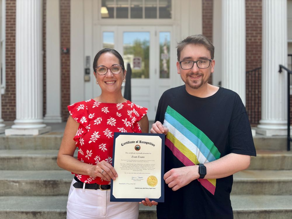 Jessica Fitzwater [left] stands in front of white columns in a red shirt with white ferns on it next to Evan Evans [right], wearing a Disability Pride Flag shirt and holding a proclamation 
