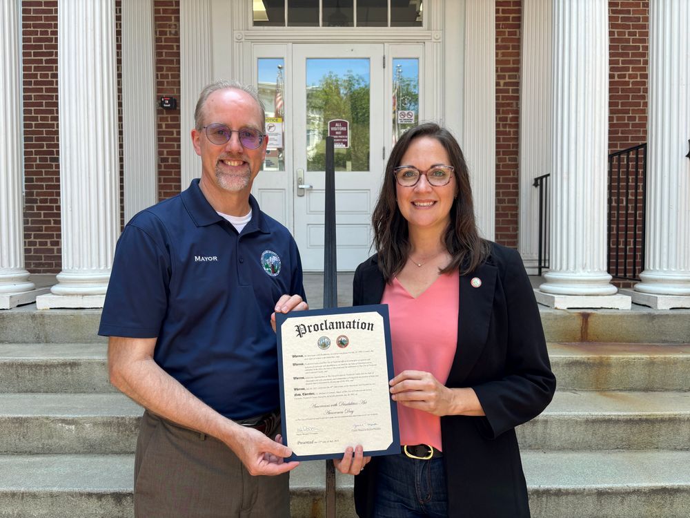 Standing in front of Winchester Hall's front doors, Mayor M. O'Connor {left} and Jessica Fitzwater, in a neon pink blouse and black blazer, on the right jointly holding a proclamation declaring July 26, 2025, as Americans with Disabilities Act Awareness Day
